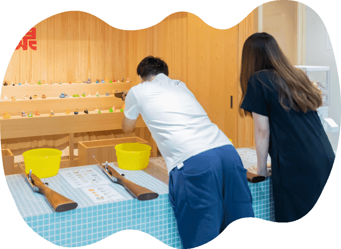 射的場で射的を楽しむ男女。男性が射的用の銃を構えて狙いを定めている。女性が後方で見守っている